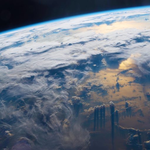 A photograph of Earth viewed from space, showing the bright blue curve of the atmosphere against the black void. Swirling white clouds cover much of the planet below, while golden sunlight reflects off the ocean surface, casting long dark shadows from towering cloud formations. Source: ISS/NASA (downloaded 24 Aug. 2018) from https://www.theguardian.com/environment/2017/aug/18/climate-scientist-clouds-climate-change-interview-kate-marvel