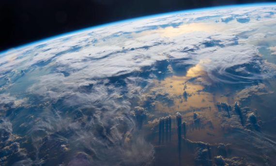 A photograph of Earth viewed from space, showing the bright blue curve of the atmosphere against the black void. Swirling white clouds cover much of the planet below, while golden sunlight reflects off the ocean surface, casting long dark shadows from towering cloud formations. Source: ISS/NASA (downloaded 24 Aug. 2018) from https://www.theguardian.com/environment/2017/aug/18/climate-scientist-clouds-climate-change-interview-kate-marvel